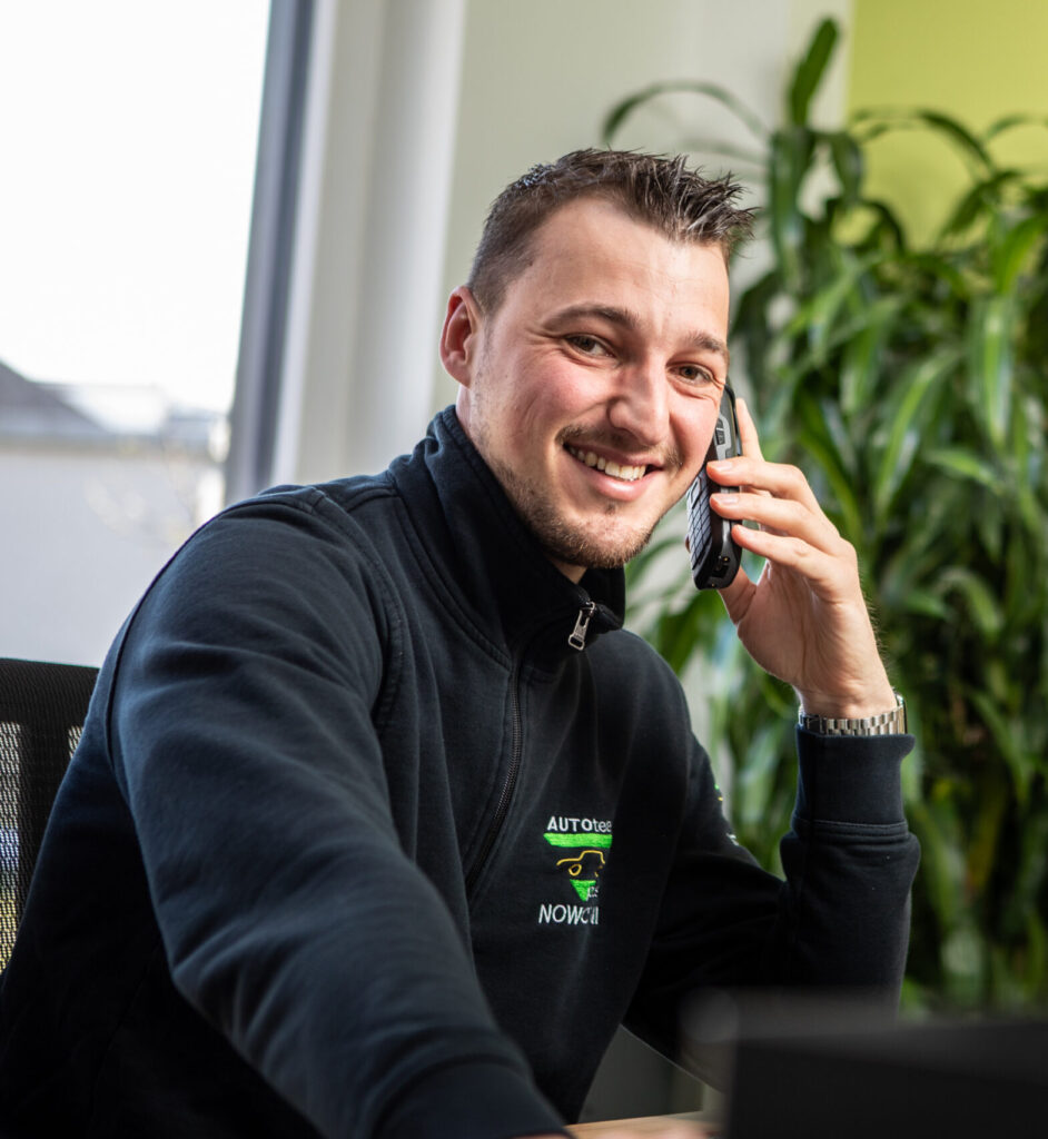 Eine Person in schwarzer Arbeitskleidung mit "AUTOTEAM plus NOWOTNI"-Logo telefoniert in einem Büro mit Pflanzen im Hintergrund.