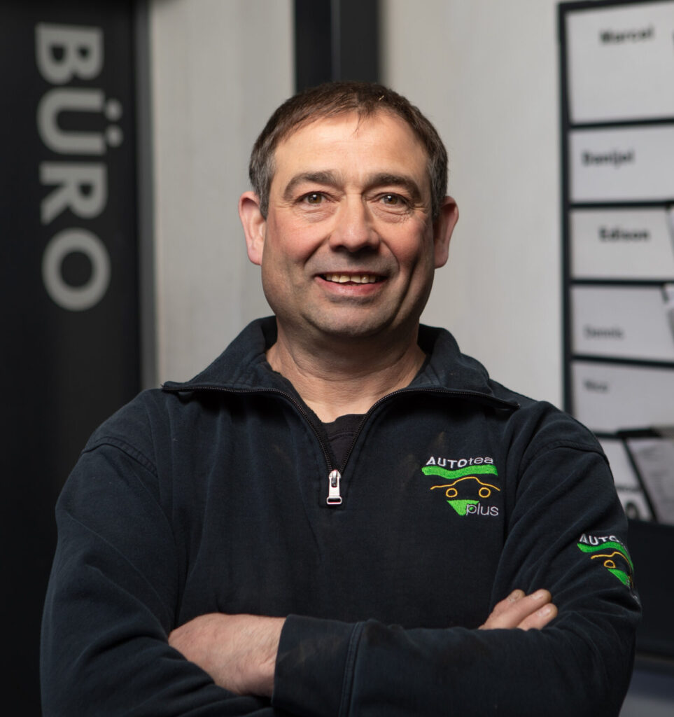 Eine Person in schwarzer Arbeitskleidung mit "AUTOTEAM plus"-Logo steht mit verschränkten Armen vor einem Bürobereich.