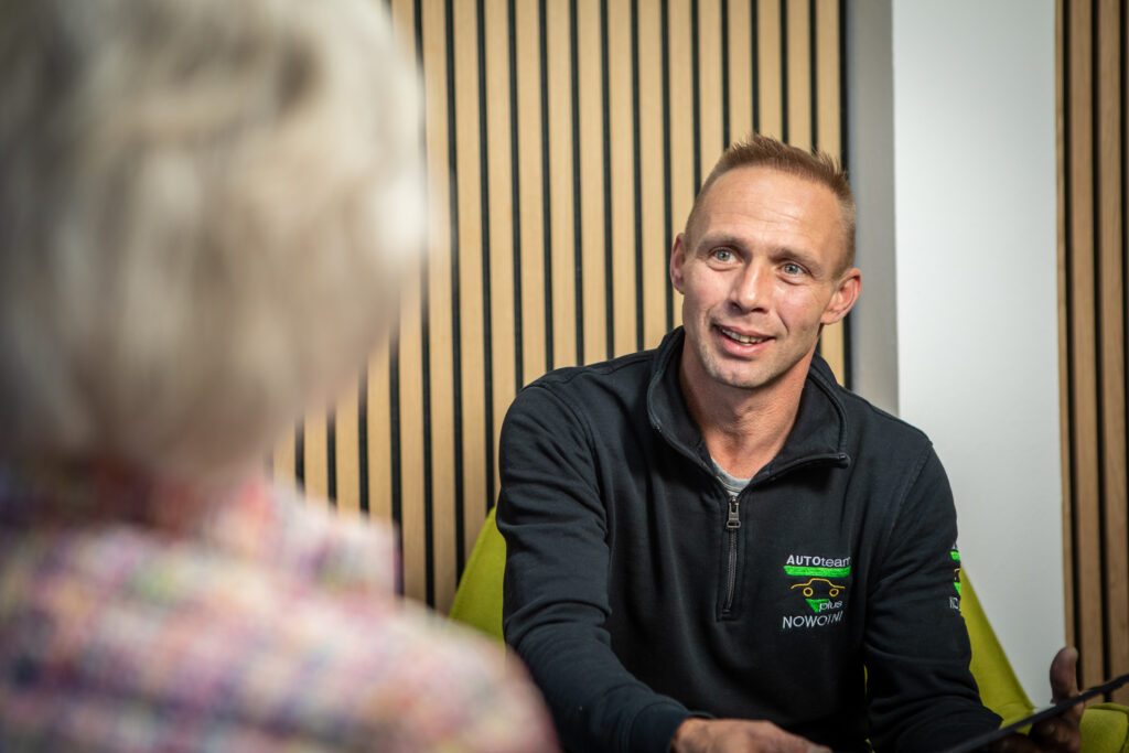 Eine Person in schwarzer Arbeitskleidung mit "AUTOTEAM plus NOWOTNI"-Logo sitzt in einem Gespräch, vor einer Wand mit Holzpaneelen.