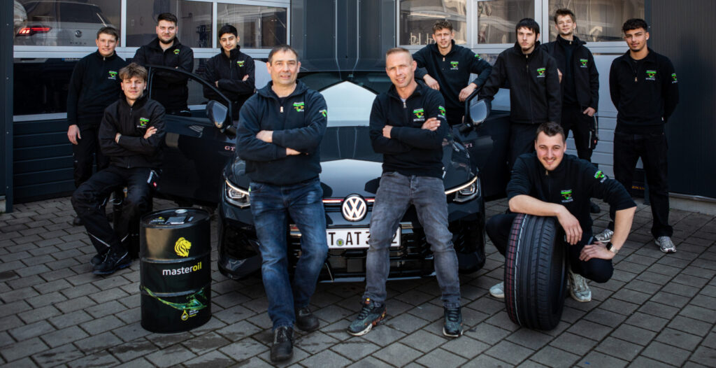 Ein Team von Mitarbeitern posiert vor einem schwarzen VW Golf GTI, begleitet von einem Reifen und einem Ölfass. Im Hintergrund ist das Werkstattgebäude zu sehen.