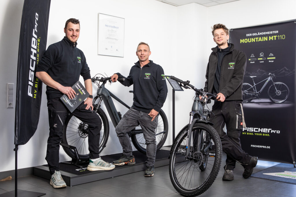 Drei Personen präsentieren moderne E-Bikes in einem Ausstellungsraum mit Bannern und Informationsmaterialien von FISCHERpro.