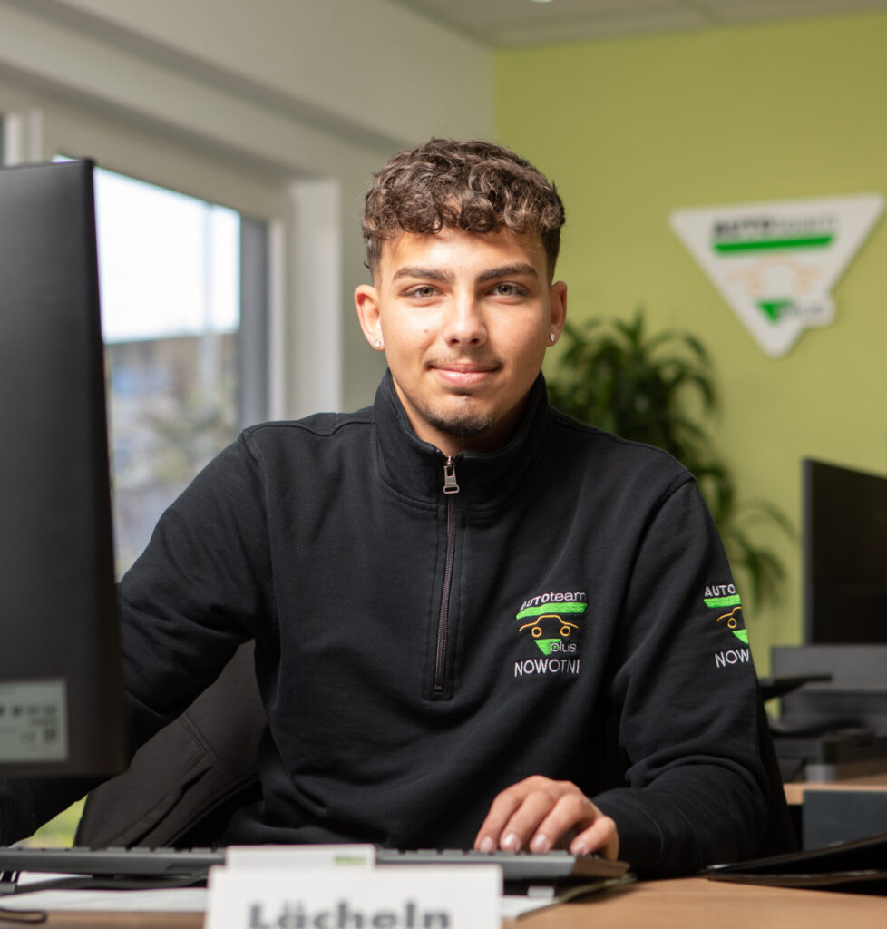 Ein Mitarbeiter in schwarzer Arbeitskleidung mit "AUTOTEAM plus NOWOTNI"-Logo arbeitet an einem Computer in einem Büro.