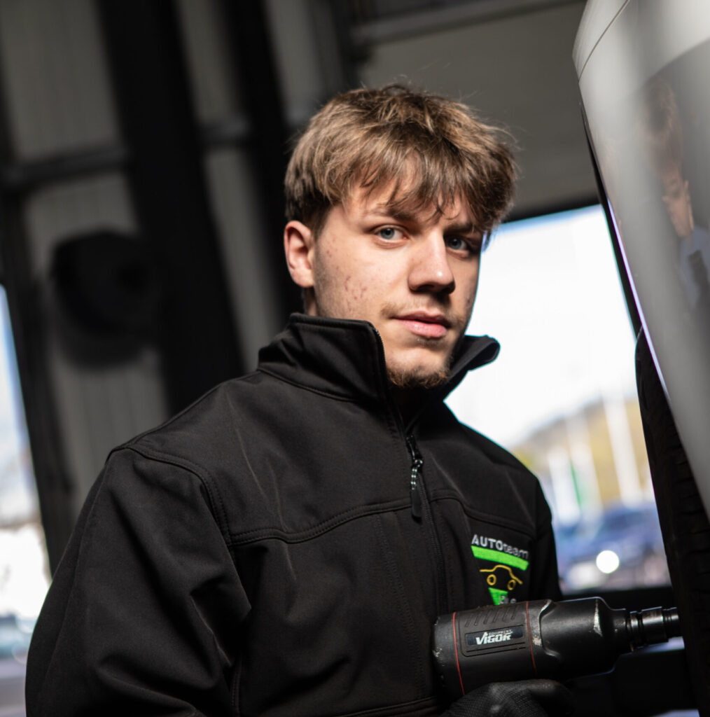 Ein Mechaniker in schwarzer Arbeitsjacke nutzt ein Druckluftwerkzeug an einem Fahrzeugreifen.