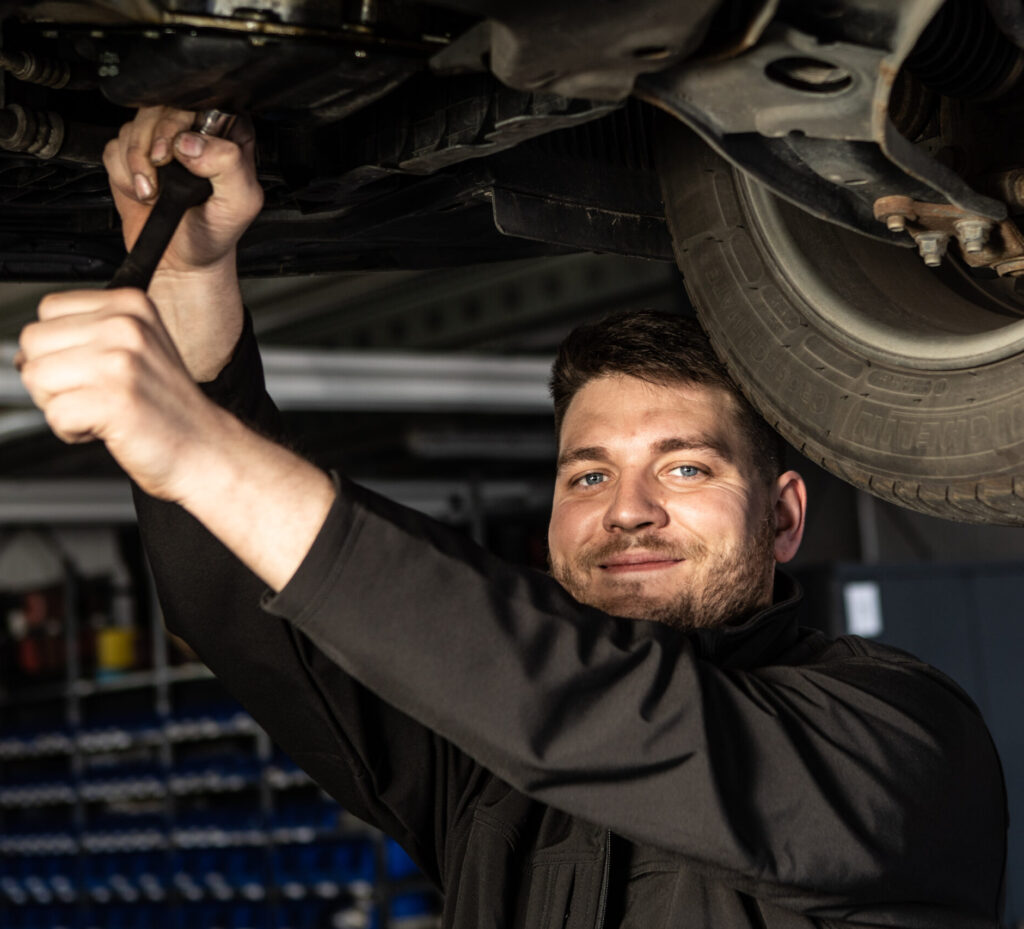 Ein Mechaniker arbeitet mit einem Werkzeug am Unterboden eines Fahrzeugs, daneben ist ein Reifen sichtbar.