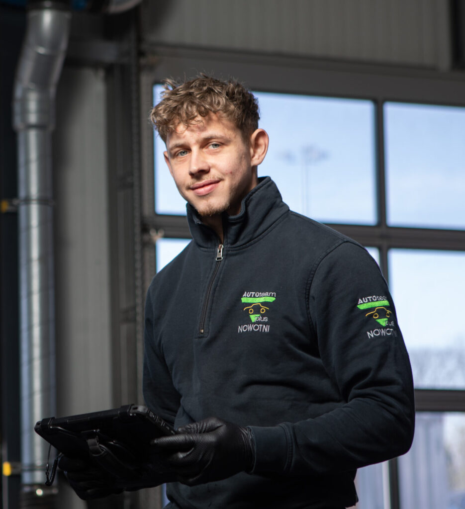 Ein Mitarbeiter in schwarzer Arbeitskleidung mit "AUTOTEAM plus NOWOTNI"-Logo hält ein Tablet in einer Werkstatt.