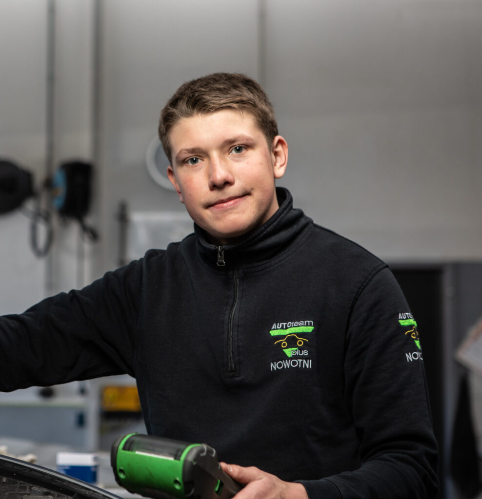 Ein Werkstattmitarbeiter in schwarzer Jacke mit dem Logo "AUTOTEAM plus NOWOTNI", hält ein grünes Werkzeug. Im Hintergrund Werkstattausstattung.