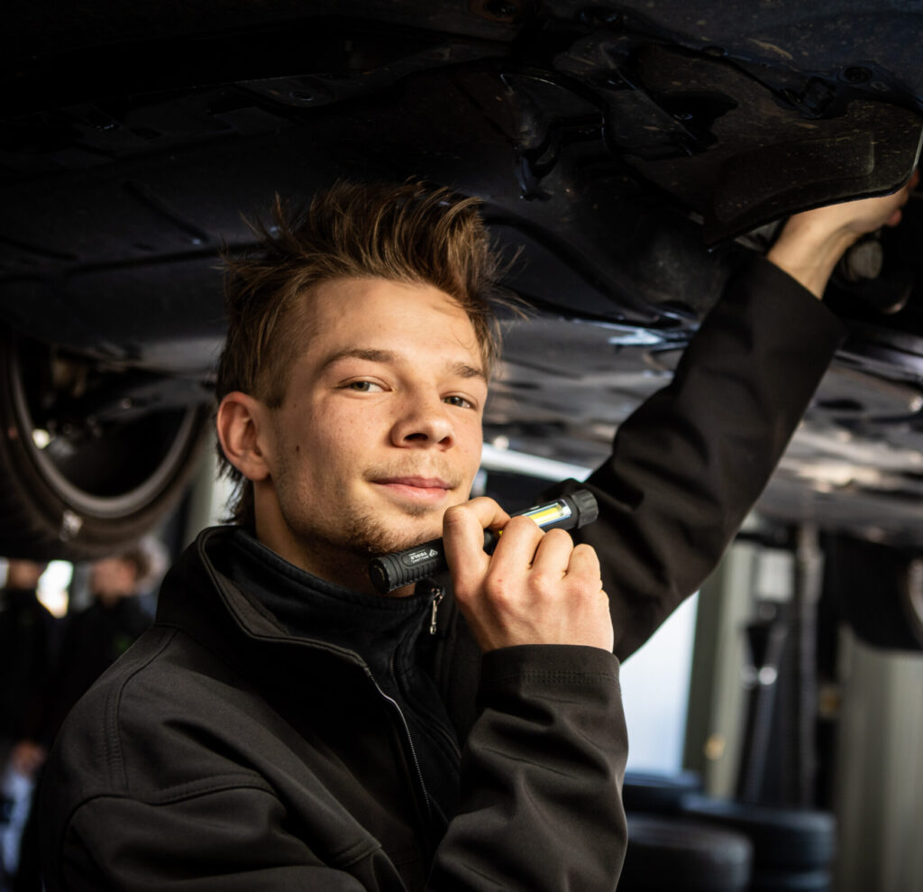 Ein Mechaniker inspiziert den Unterboden eines Fahrzeugs mit einer kleinen Lampe in der Hand.